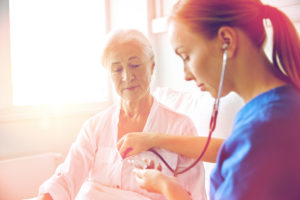 medicine, age, support, health care and people concept - doctor or nurse with stethoscope visiting senior woman and checking her heartbeat at hospital ward