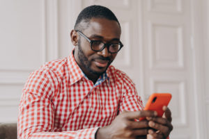 Cheerful millennial biracial man using smartphone, sitting at desk , young Afro american guy carefully looking on phone screen while typing urgent message, chatting online or searching in internet