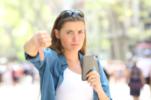 Annoyed woman holding a smart phone looking at camera with thumbs down in the street