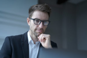 Focused German man family lawyer in formal wear thoughtfully looking at computer monitor while distantly working from modern home office carefully reading clients' prenuptial agreement for signature