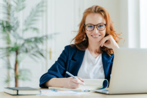 Happy successful female writes checklist in notepad, works on creative ideas for startup project, searches necessary information on laptop computer, poses at coworking space, works freelance